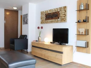 A modern living room with a flat-screen TV, a wooden console, a black leather sofa, and a decorative wall piece.