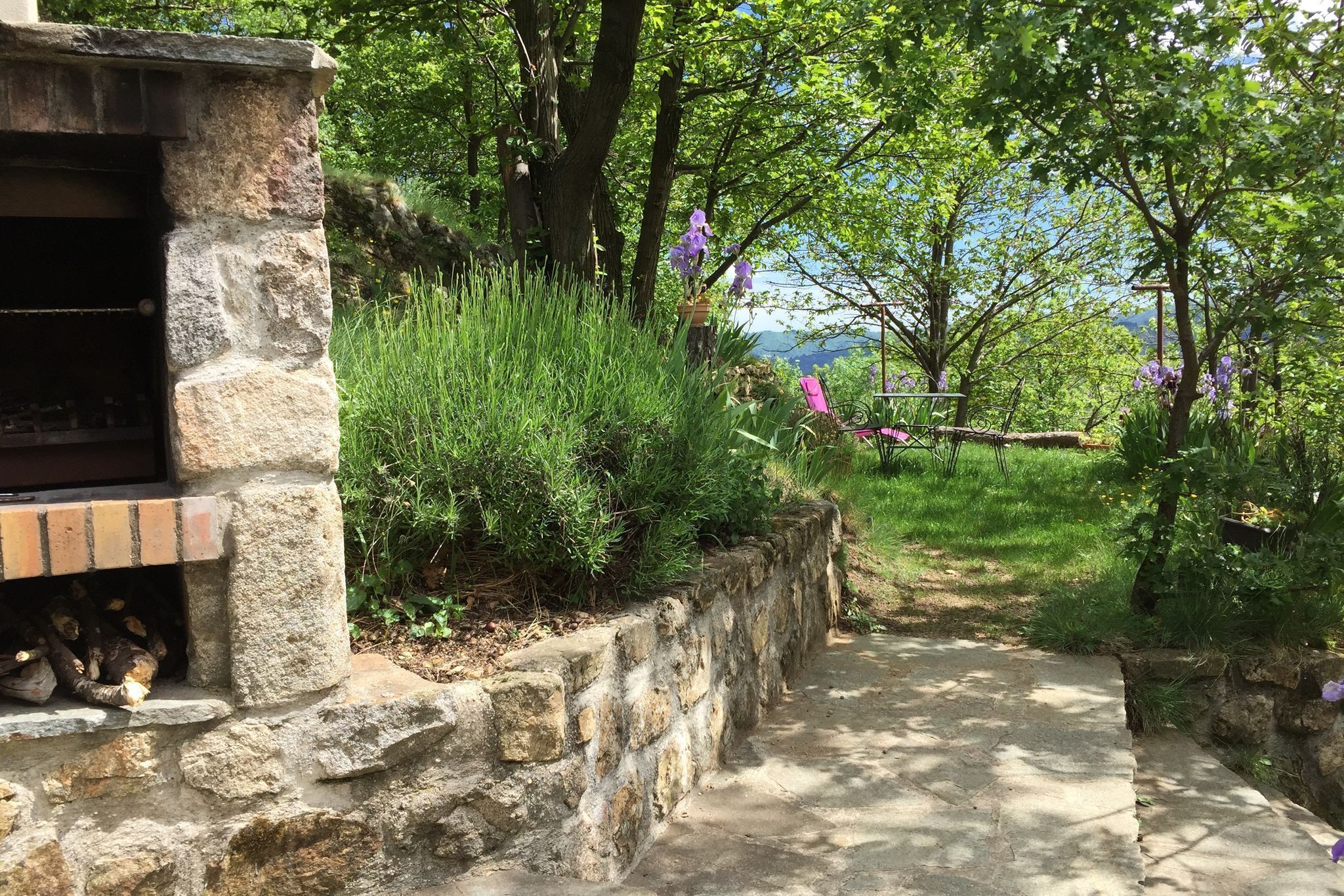 Un espace extérieur aménagé en pierre, doté d’un barbecue et entouré d’une végétation luxuriante et de plantes en fleurs.