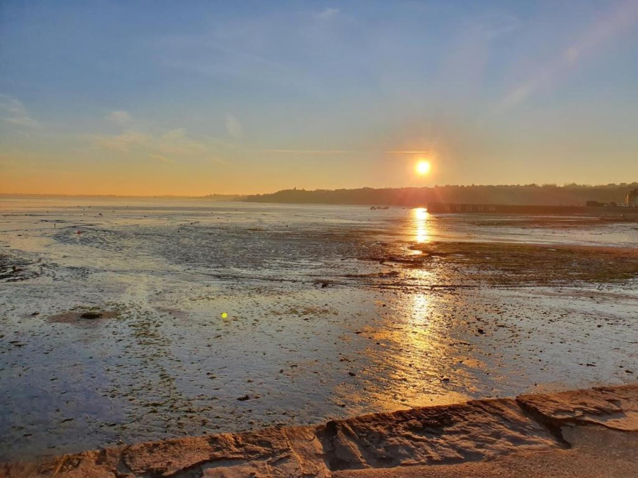 A beach at sunset with a clear sky and calm waters reflecting the sun.