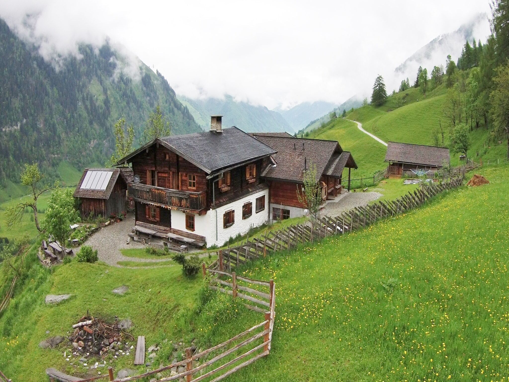 A traditional wooden house with a sloped roof, surrounded by lush green fields and mountains.