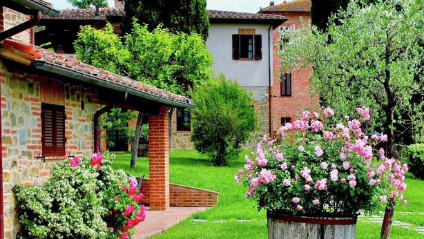 A rustic stone building with a tiled roof, a brick pathway, and a flower bed with pink roses.