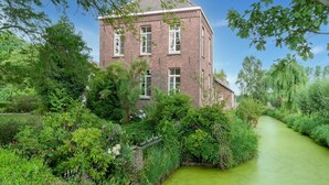 A brick building with multiple windows, surrounded by lush greenery and a narrow water channel.