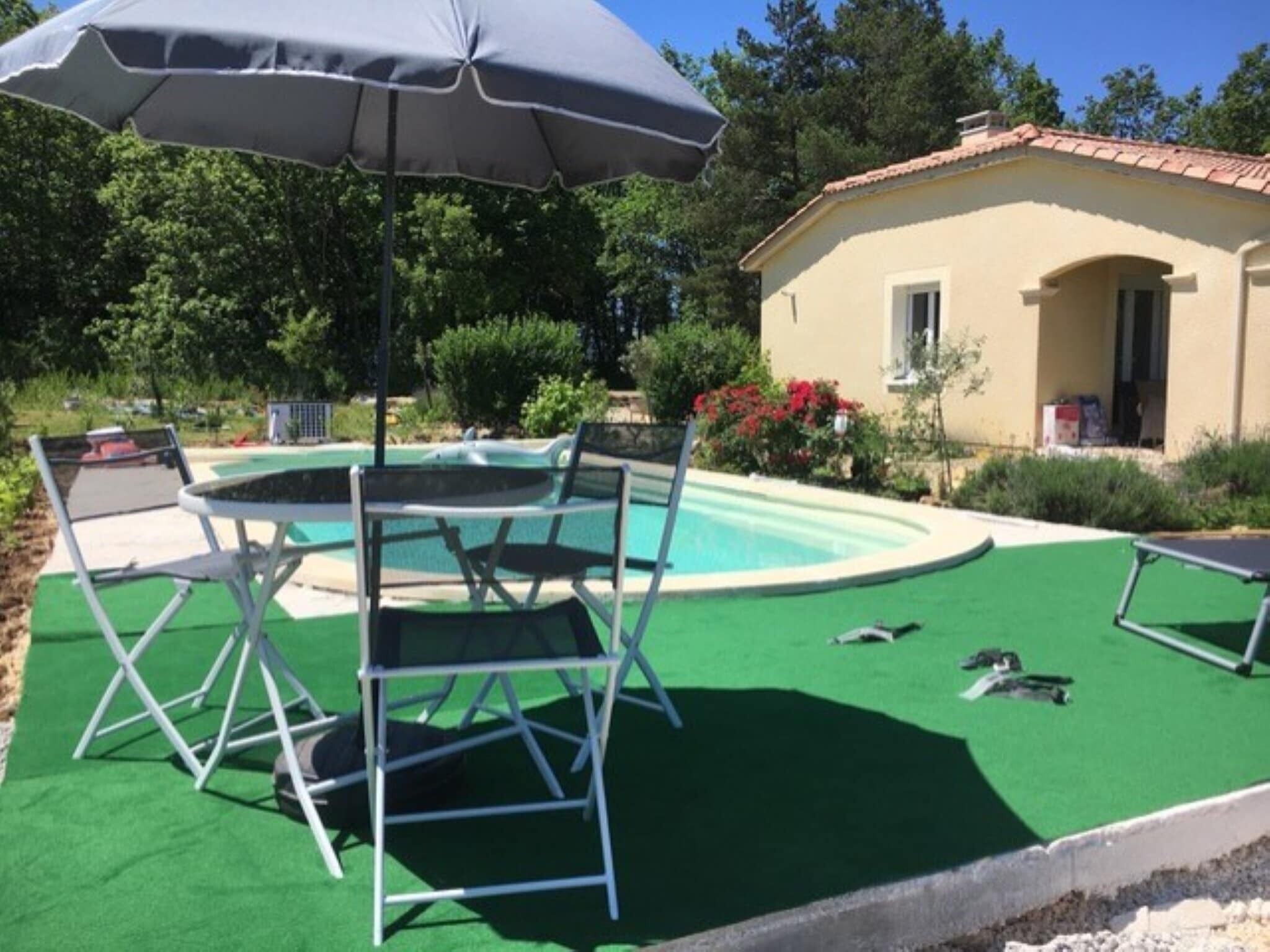 A pool area with a table and chairs, an umbrella, and a small building in the background.