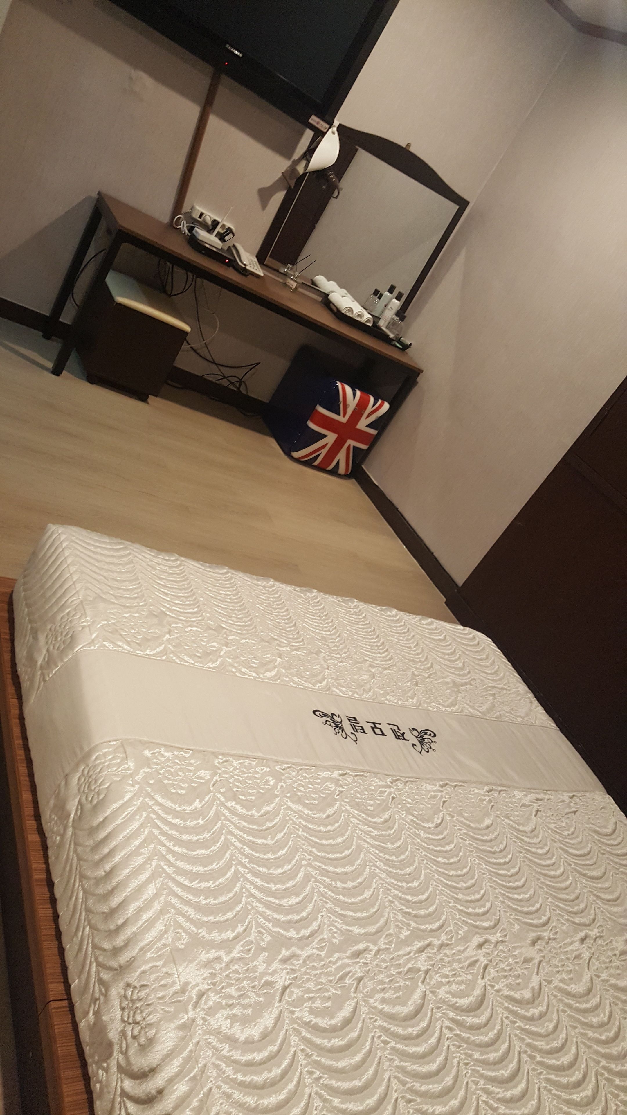 A neatly made bed with a decorative headboard, a desk with a computer and a phone, and a wall-mounted TV.
