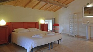 Ein Schlafzimmer mit Holzbalkendecke, einem Bett mit weißer Bettwäsche, einem Nachttisch mit Lampe, einem Kleiderschrank und einem Badezimmer mit Waschbecken und Spiegel.