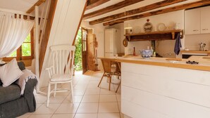 A cozy kitchen with wooden beams, a dining area with a white chair, and a kitchen counter with various items.