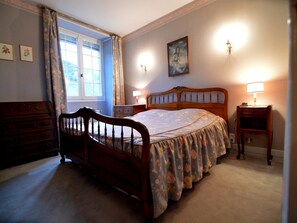 Een slaapkamer met een houten bed, een dressoir, een nachtkastje met lamp en een raam met gordijnen.