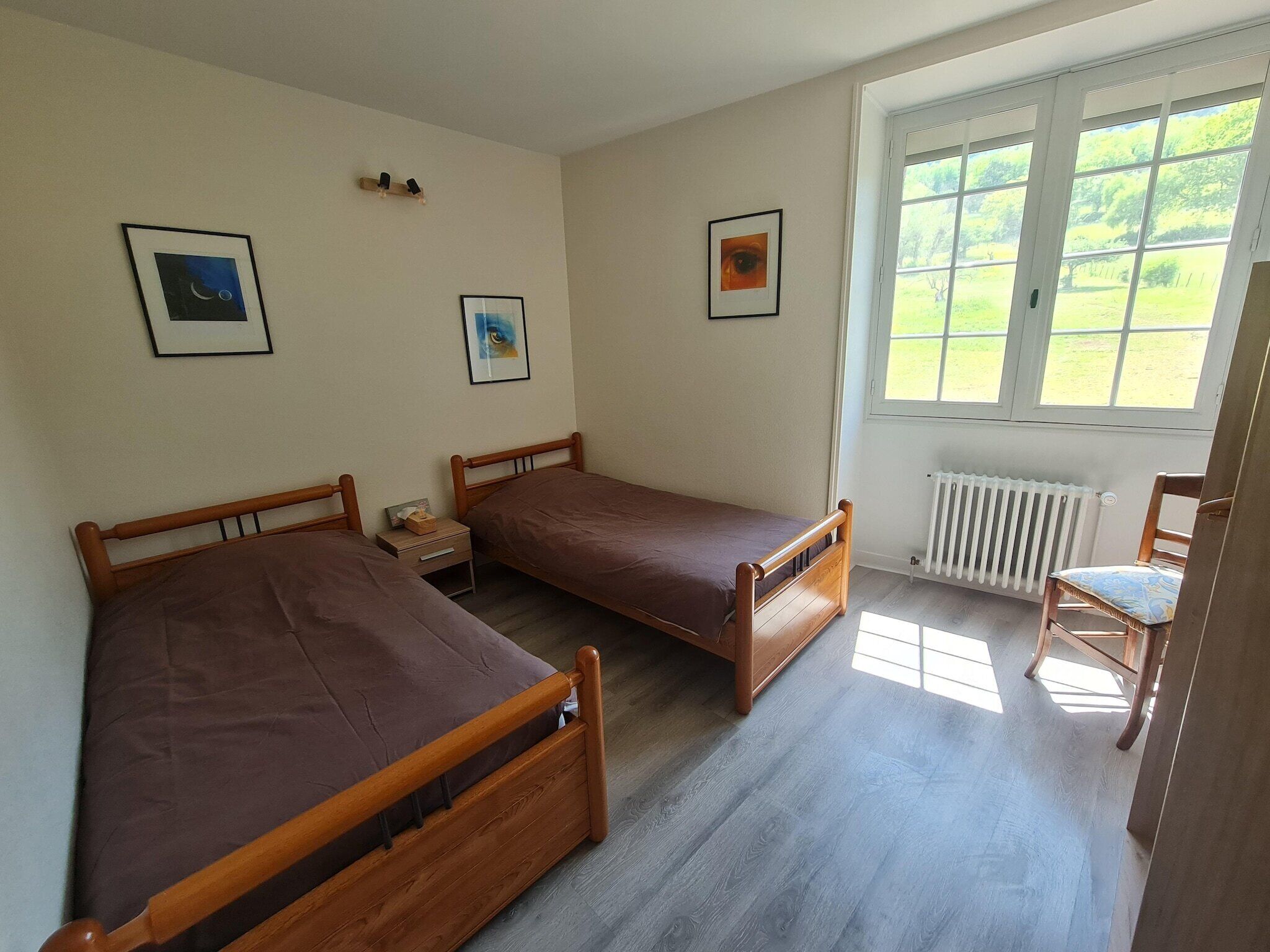 A bedroom with two wooden beds, a small bedside table, a chair, and a window with a view of greenery.