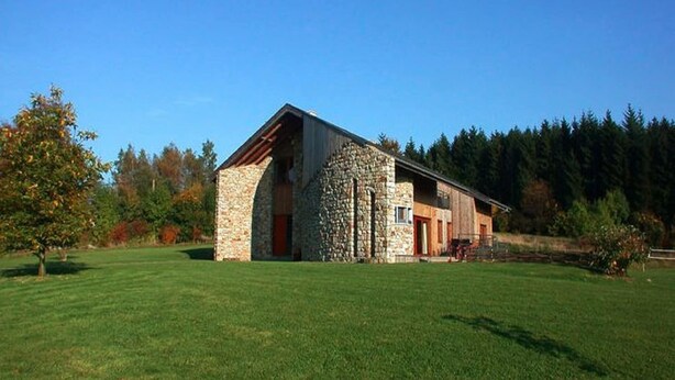 Edifici de pedra amb teulada inclinada, situat sobre un camp herbós amb arbres al fons.