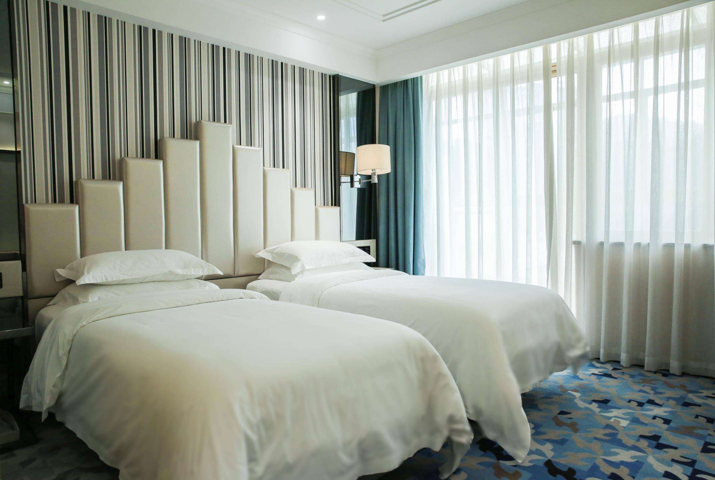 Habitación de hotel con dos camas, un muro a rayas, una lámpara y un ventanal grande con cortinas.