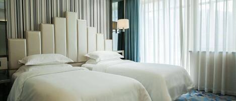 Habitación de hotel con dos camas, un muro a rayas, una lámpara y un ventanal grande con cortinas.