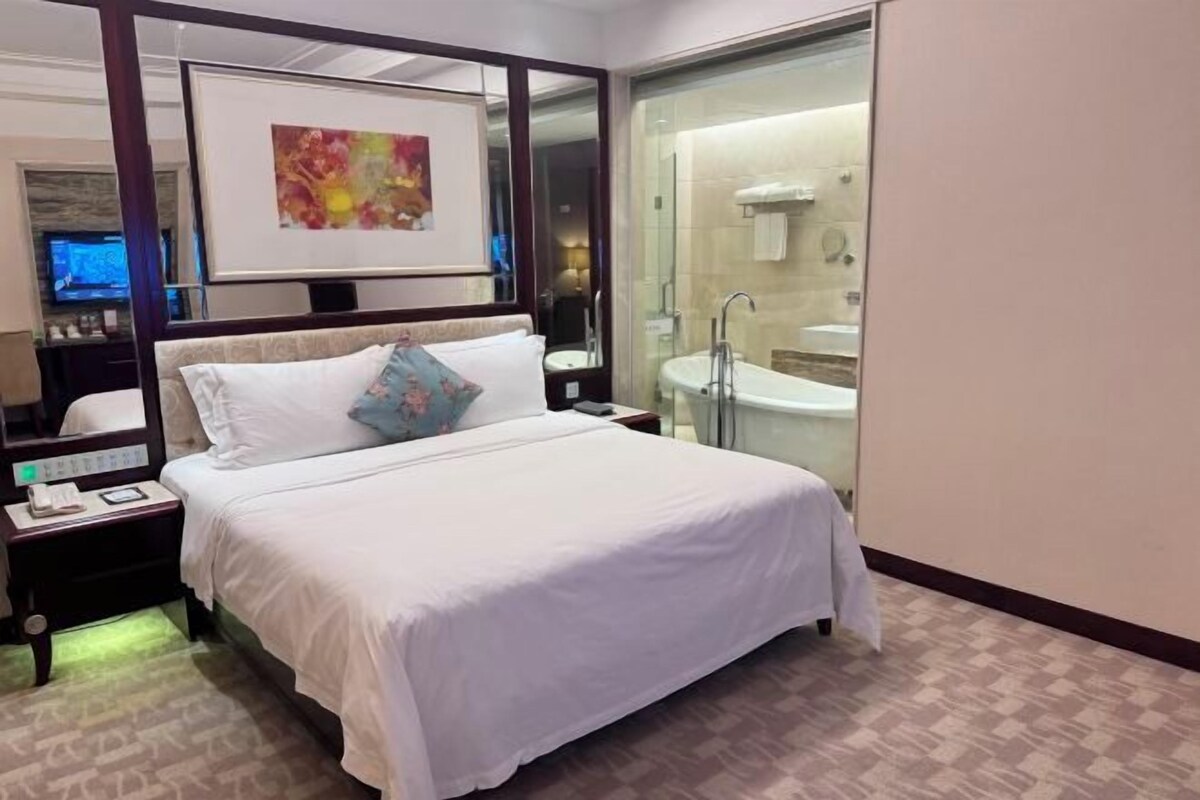 Una habitación de hotel con una cama grande, una mesita de noche con teléfono, un espejo, un cuadro en la pared y una zona de ducha con mampara de vidrio.