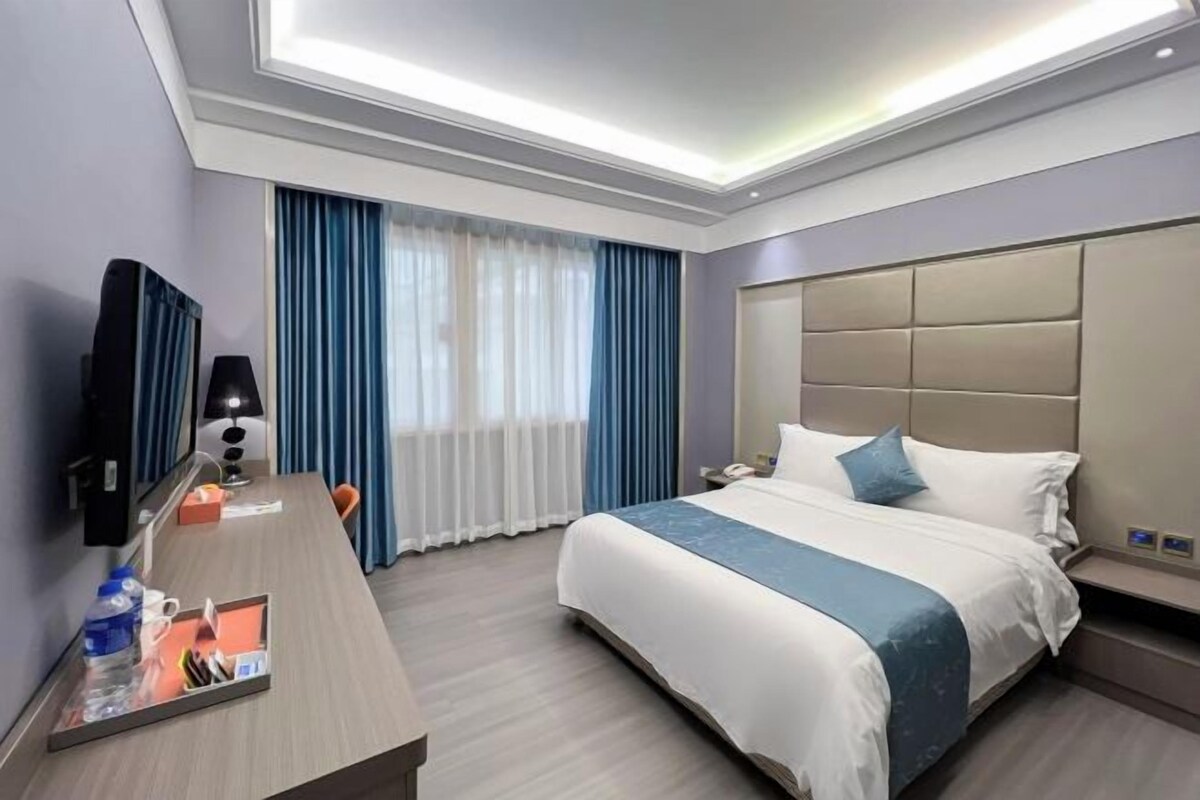 Una habitación de hotel con una cama grande, un escritorio con lámpara, un televisor y una ventana con cortinas.
