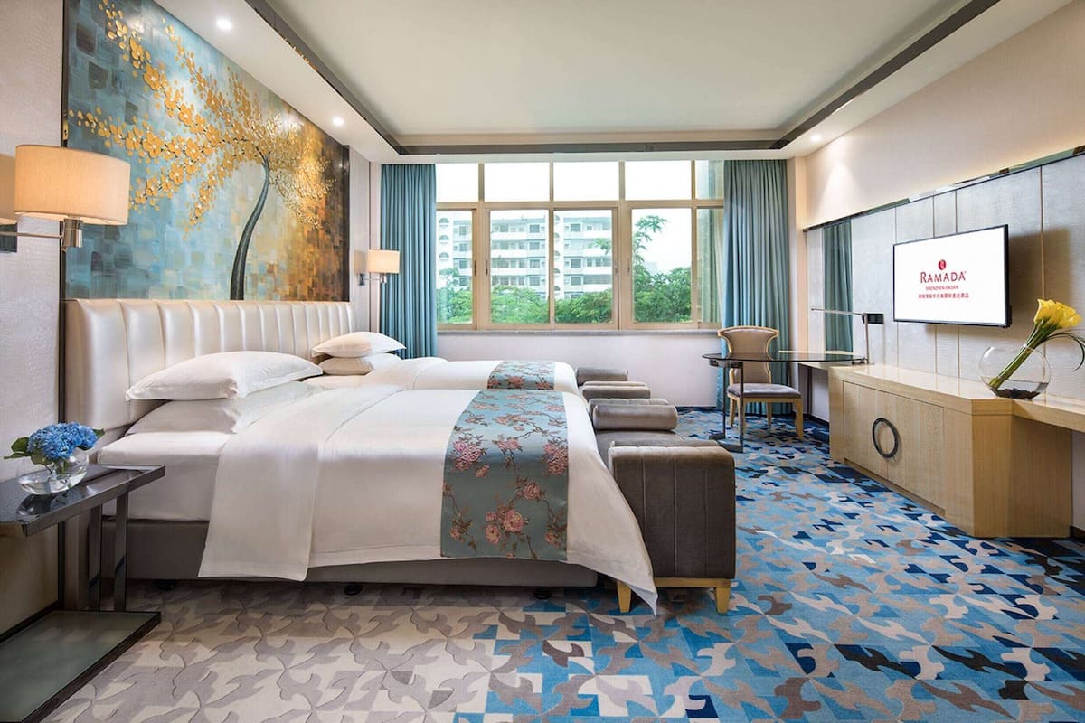 A hotel room with a large bed, a desk, a chair, a TV, and a view of buildings through the window.