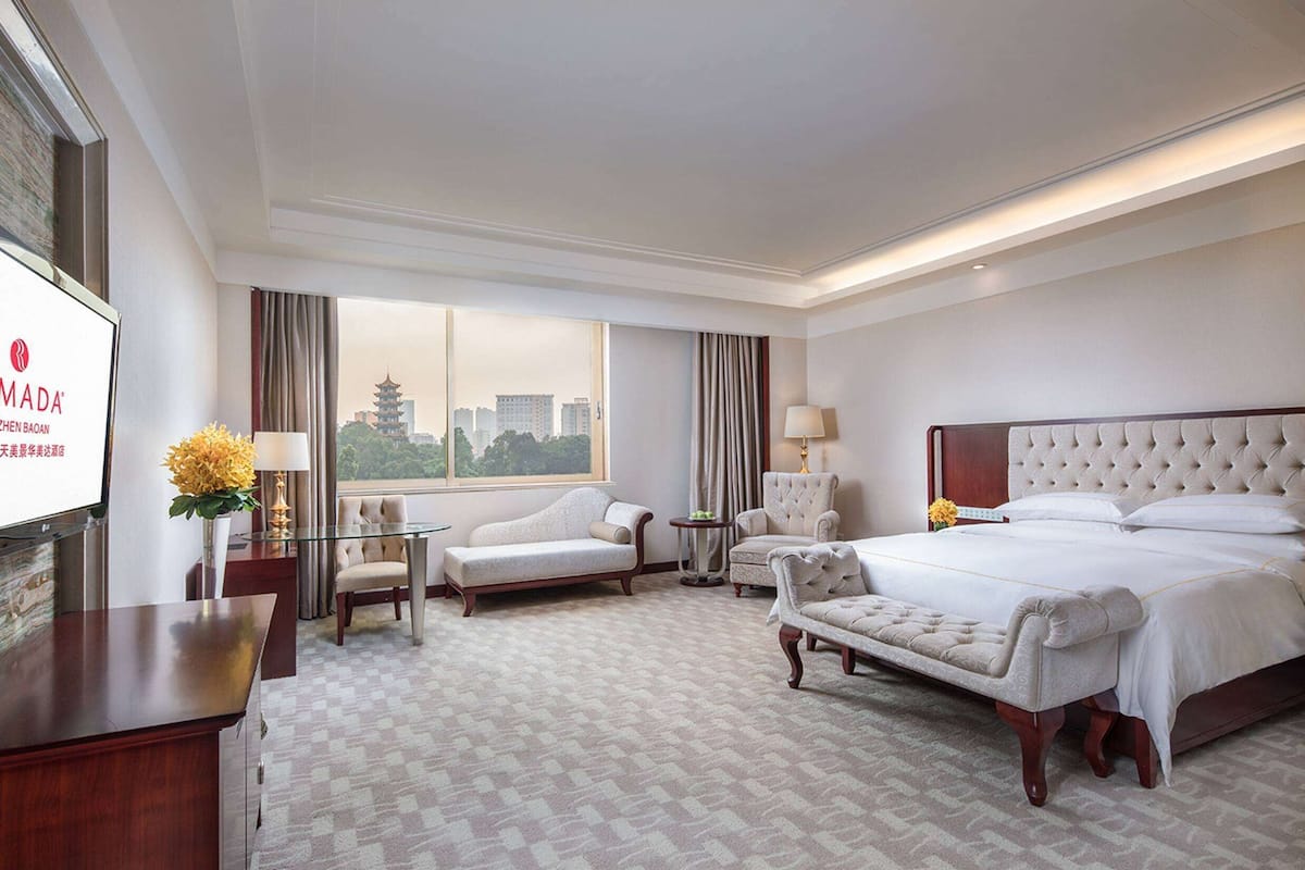 A hotel room with a large bed, a sofa, a chair, a desk, and a TV screen displaying the hotel's name.
