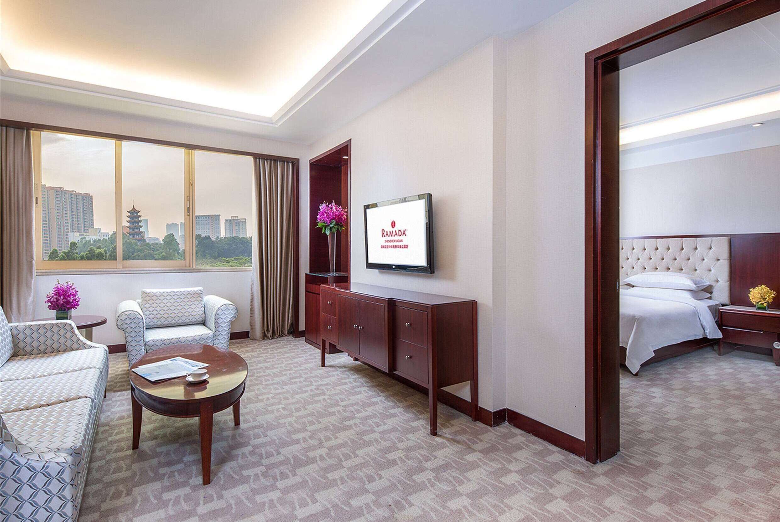 A hotel room with a sofa, a coffee table, a TV, a bed, and a view of the cityscape.