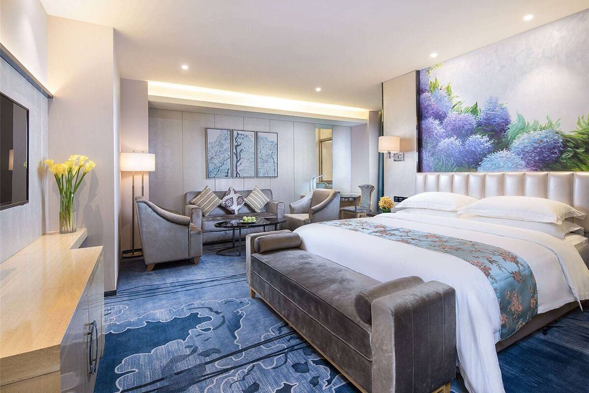 A modern hotel room with a large bed, a seating area with chairs, a desk, and a painting on the wall.