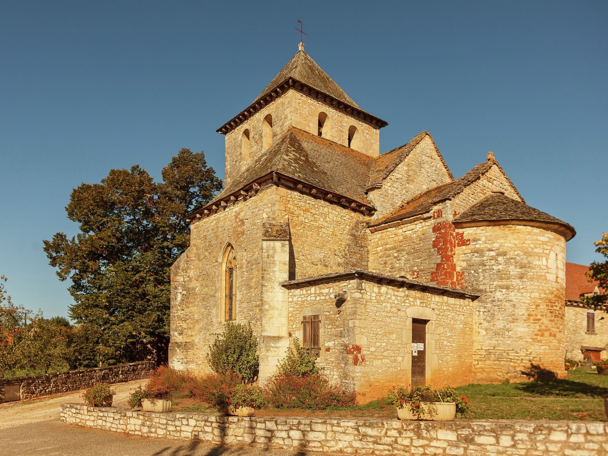Una iglesia de piedra histórica con un campanario y una torre redonda, rodeada por un muro de piedra y follaje.
