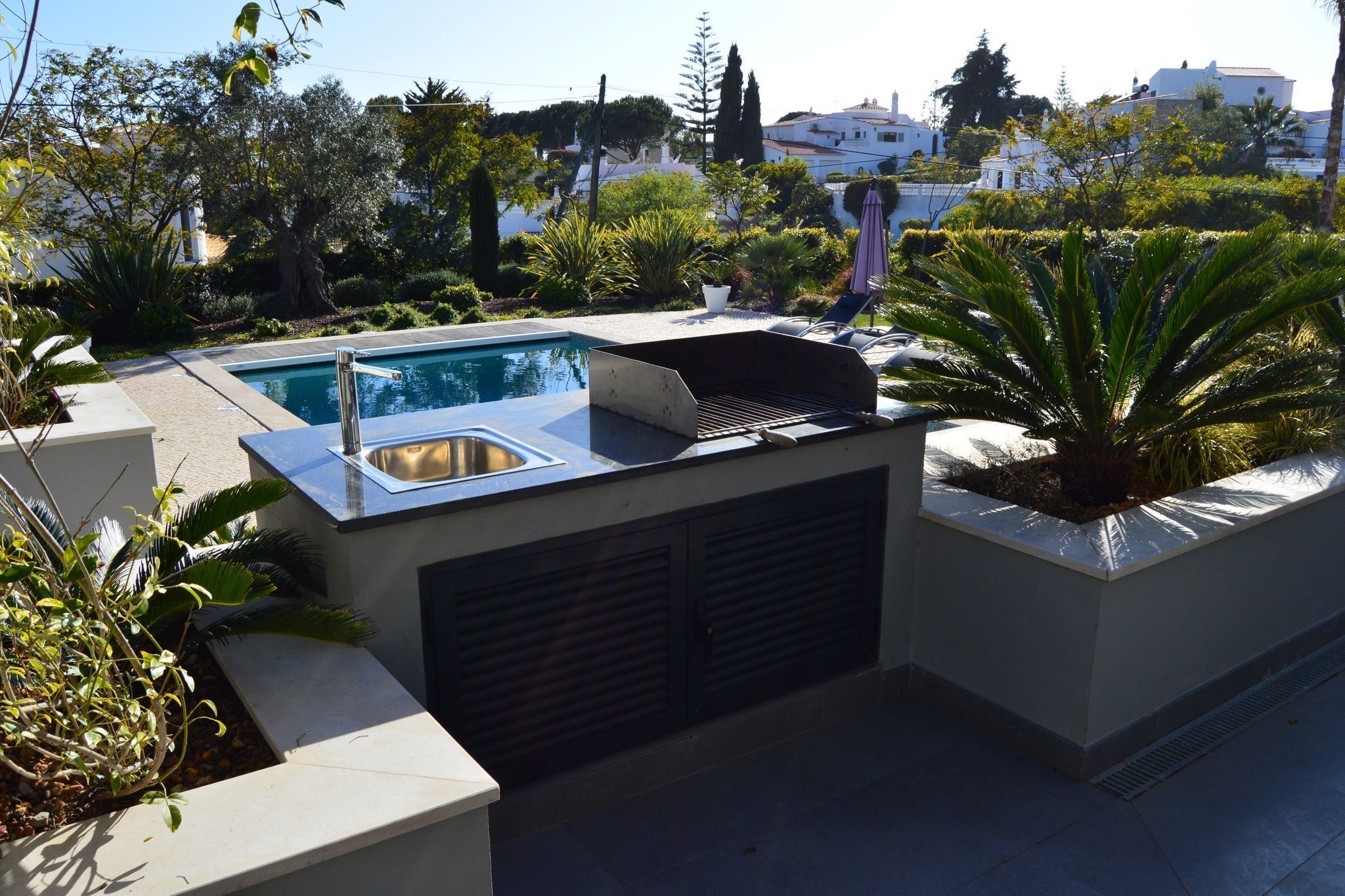 A rooftop pool area with a built-in grill and a sink, surrounded by potted plants and overlooking a residential neighborhood.