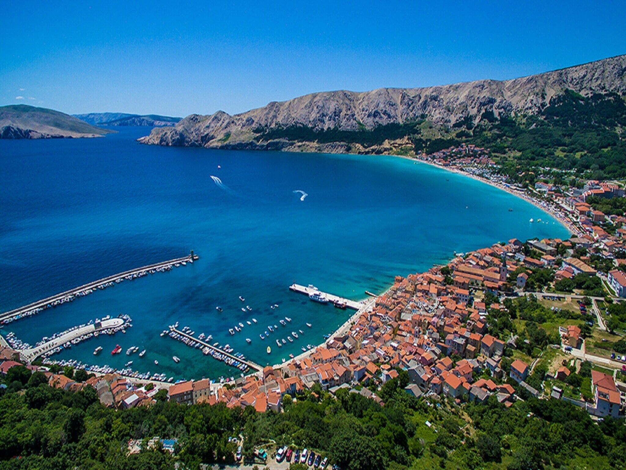 An aerial view of a coastal town with a marina, boats, and buildings with red-tiled roofs.