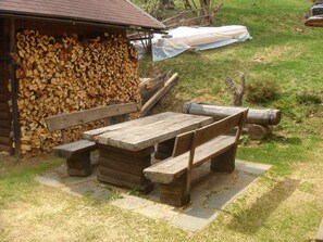 Un espace extérieur aménagé dans un style rustique, avec une table et des bancs en bois, une pile de bois de chauffage et une structure couverte.