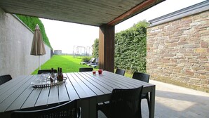 Une terrasse extérieure avec une table dressée pour deux personnes, un plateau en métal avec des verres et des bouteilles, et une vue sur une pelouse verdoyante et un mur de pierre.