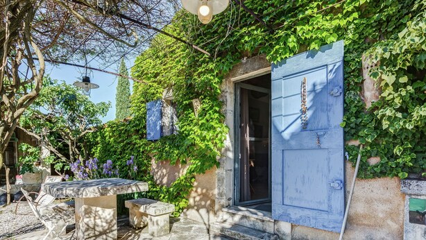 A rustic outdoor seating area with stone tables and benches, surrounded by lush greenery and a blue door. - Cottage in Oppède amidst Vineyards (Oppède)