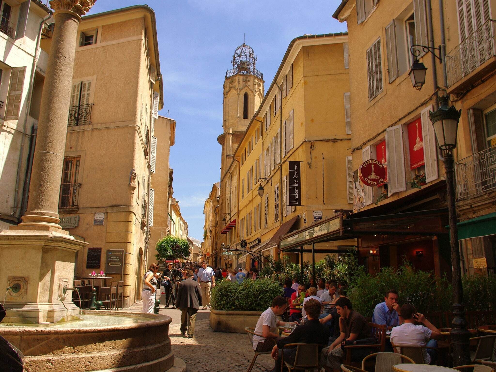 A narrow street with buildings on either side, a fountain in the center, and people sitting at outdoor tables.
