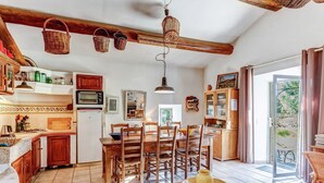 A cozy kitchen with wooden cabinets, a dining table, and chairs. There is a large window with a view of greenery outside. - Cottage in Oppède amidst Vineyards (Oppède)