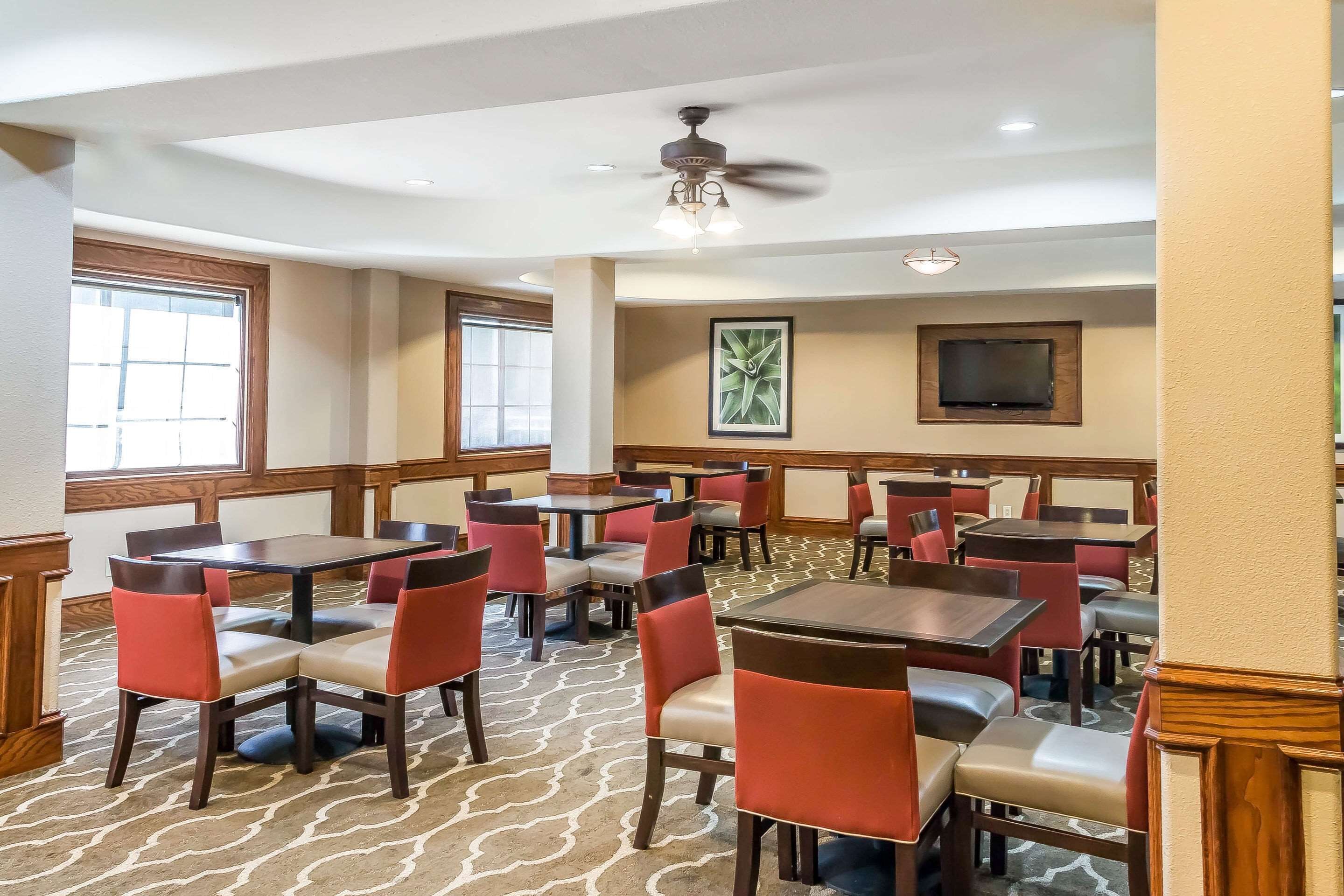 A hotel dining area with multiple tables and chairs, ceiling fans, and a television on the wall.