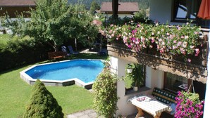 A swimming pool surrounded by lush greenery, a wooden deck, and a balcony with hanging flowers.