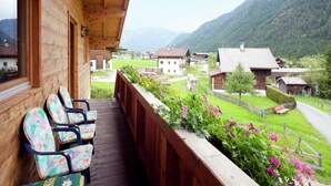 A wooden balcony with chairs and a flower box overlooking a village and mountains.