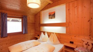 A wooden cabin room with a bed, a small table, a chair, and a window with curtains.