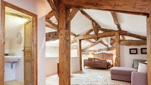 A rustic attic bedroom with exposed wooden beams, a wooden door leading to a bathroom, a bed with a white bedspread, and a purple sofa.