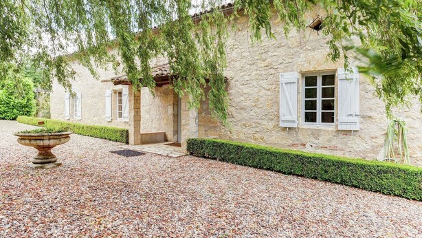 A rustic stone building with a gravel driveway, a well-maintained hedge, and a large stone planter.
