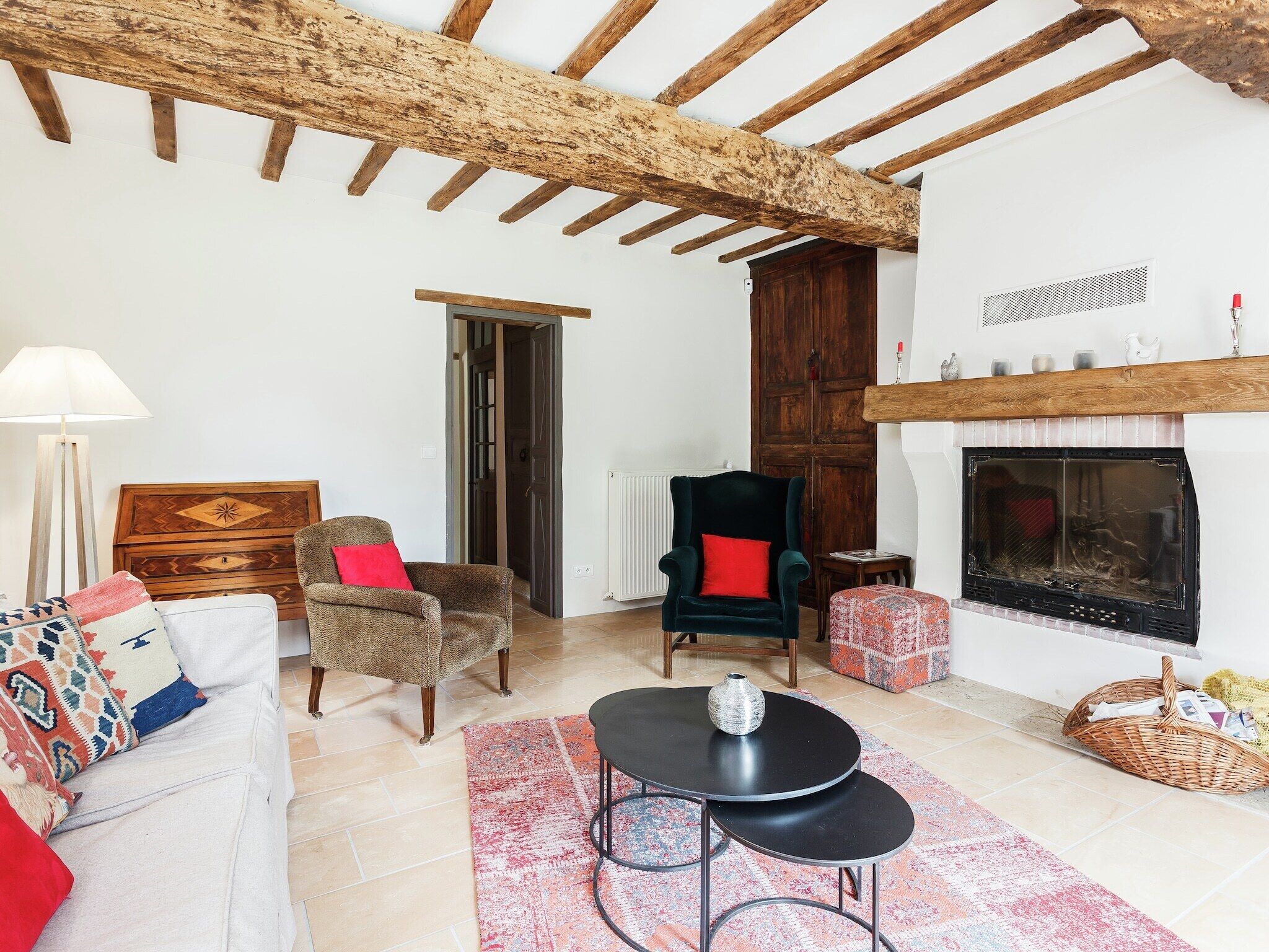 A cozy living room with a fireplace, wooden beams, a sofa with patterned cushions, a chair with a red cushion, and a small table with a vase.