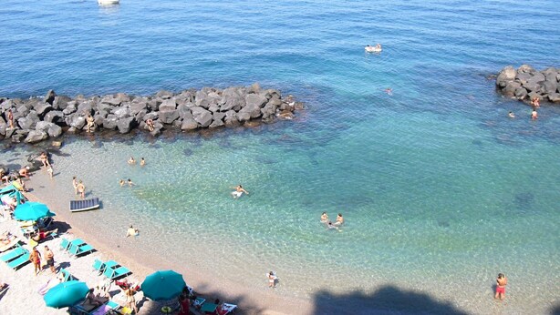 Una playa con agua cristalina, un rompeolas rocoso y gente nadando y tomando el sol.
