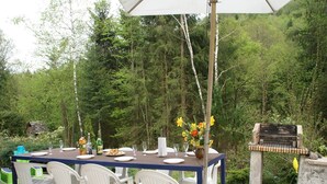 Un espacio de comedor al aire libre con una mesa dispuesta para seis personas, rodeada de sillas de plástico blanco, un banco de madera y una parrilla.