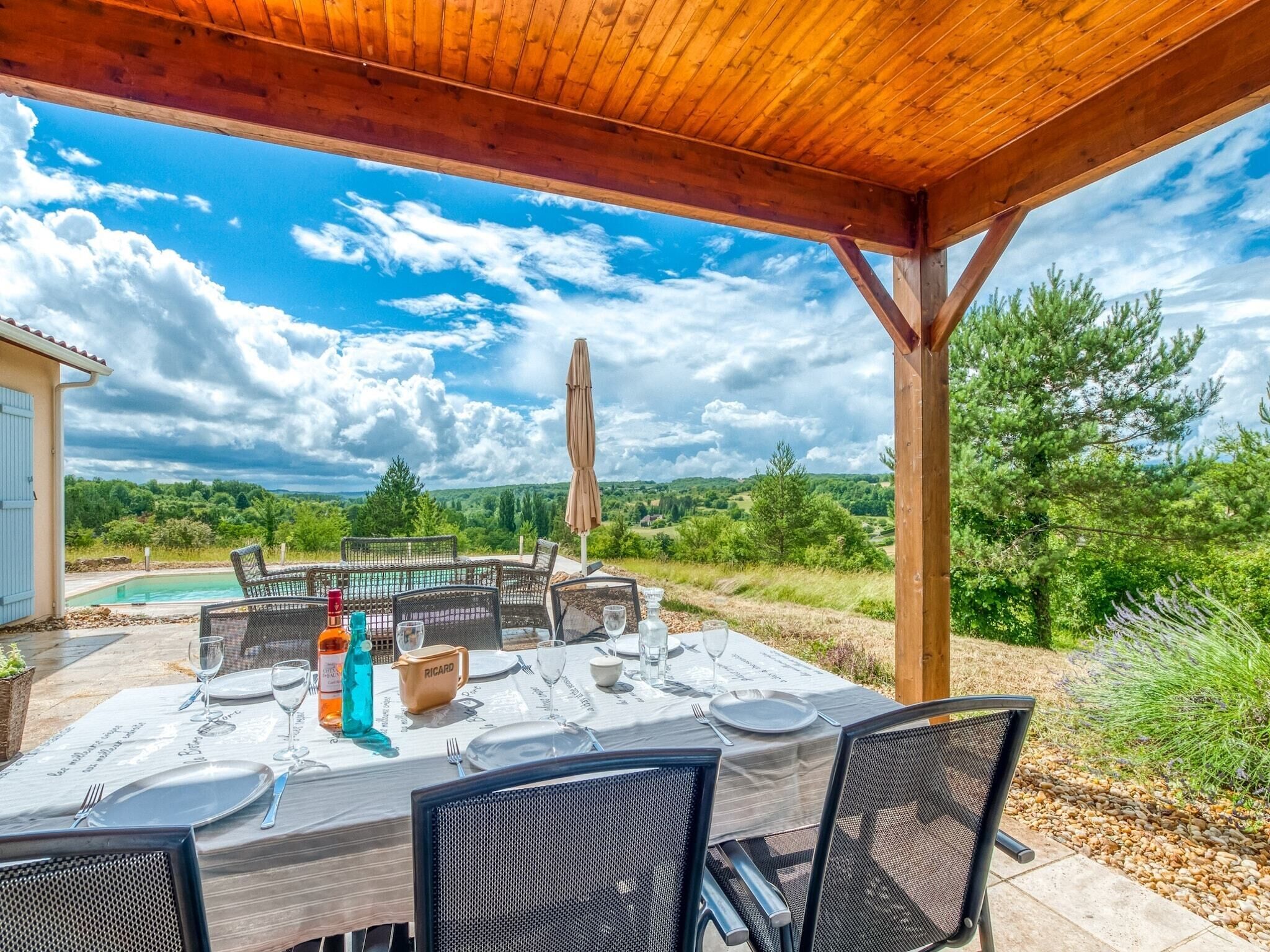 Un espace de restauration en plein air avec une table dressée pour quatre personnes, entourée de chaises, sous une pergola en bois offrant une vue sur une piscine et un paysage luxuriant.