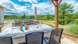 Een buiten eethoek met een tafel voor vier personen, omringd door stoelen, onder een houten pergola met uitzicht op een zwembad en een weelderig landschap.