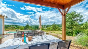 Un espace de restauration en plein air avec une table dressée pour quatre personnes, entourée de chaises, sous une pergola en bois offrant une vue sur une piscine et un paysage luxuriant.
