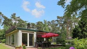 Een klein, gelijkvloers huis met een patio, compleet met een eettafel, stoelen, een rode parasol en potplanten.
