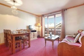 A room with a dining table, chairs, a sofa, a TV, and a window with a view of a building. - Apartment in Le Village with Balcony (Notre Dame de Bellecombe)