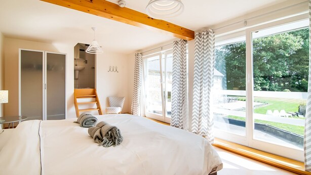 A bedroom with a large bed, a wooden ceiling beam, a window with curtains, and a view of the outdoors.