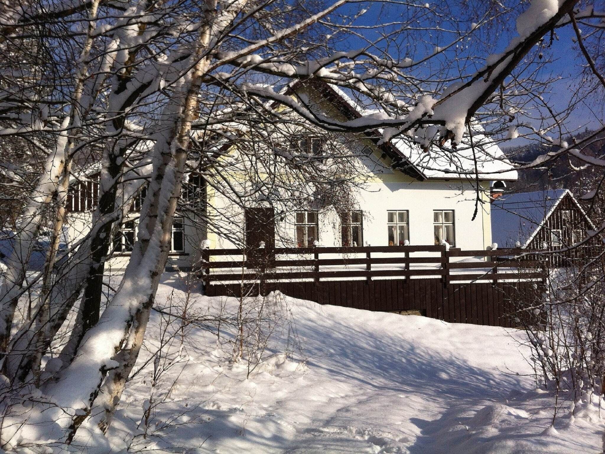 A snow-covered house with a wooden fence, surrounded by bare trees and a clear sky.
