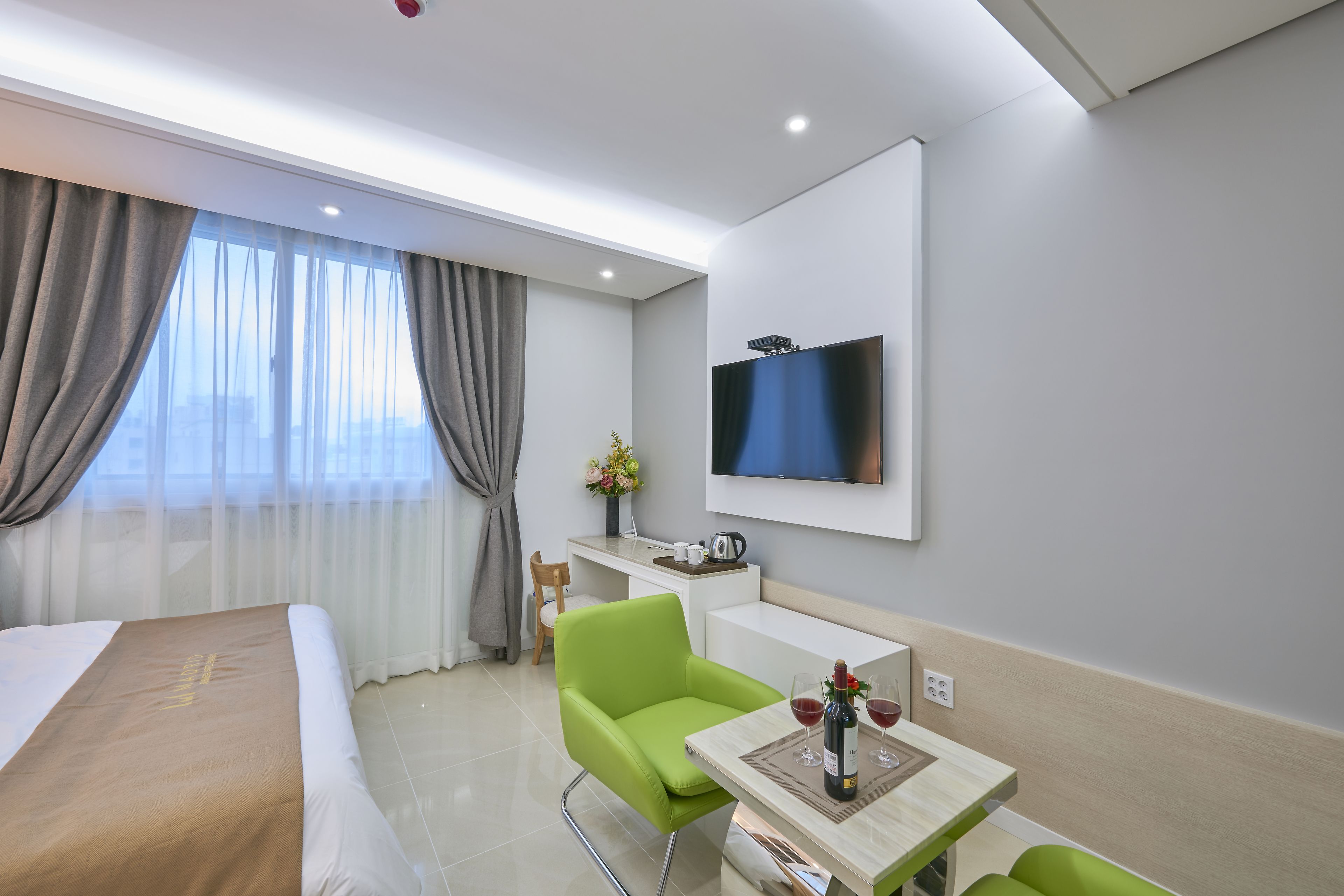 A modern hotel room with a bed, a desk, a green chair, a TV, and a small table with a bottle and glasses.