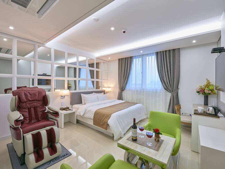 A modern hotel room with a bed, a massage chair, a dining area, and a TV.