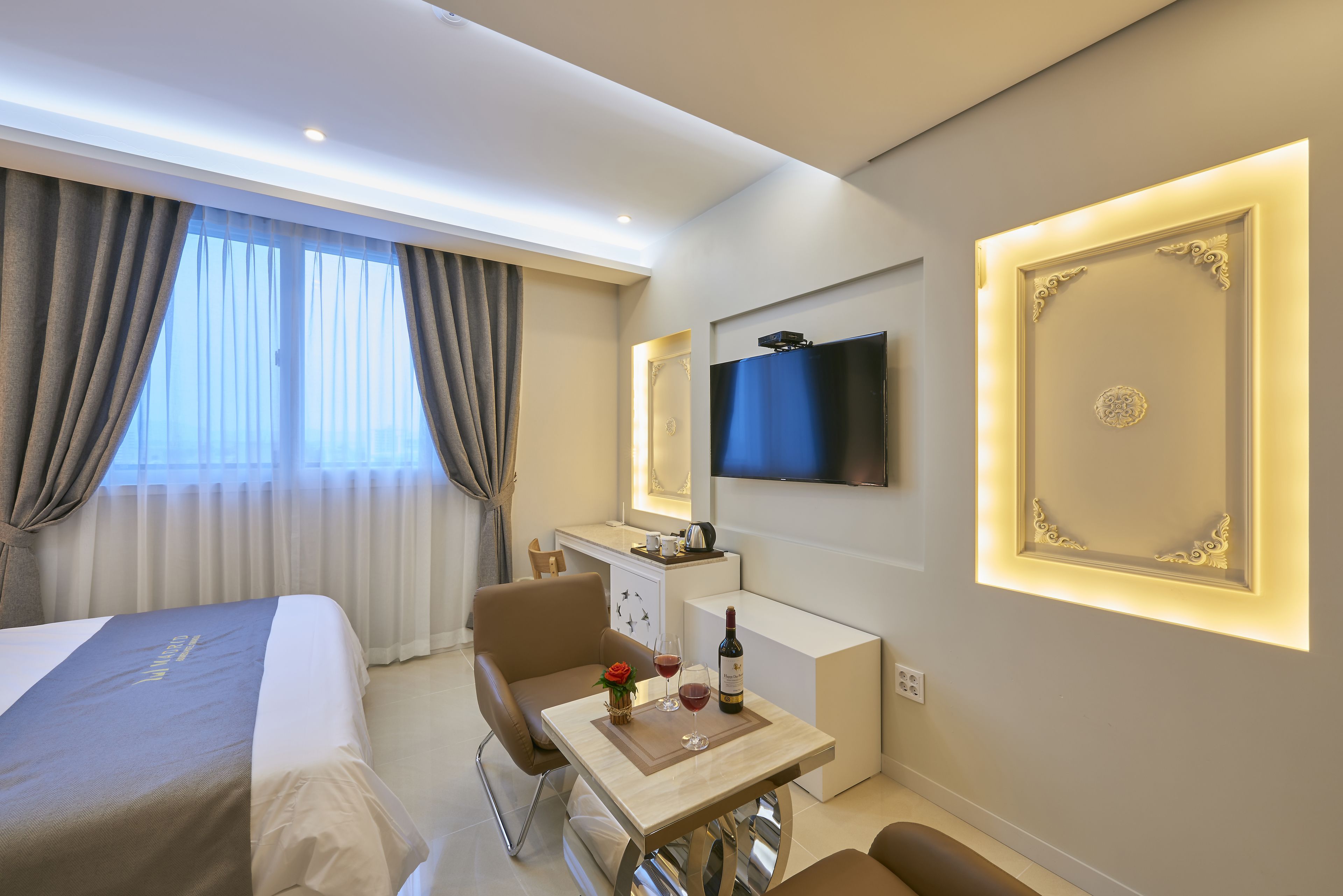 A hotel room with a bed, a desk with a chair, a television mounted on the wall, and a small table with a bottle and glasses.