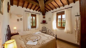 A bedroom with a wooden ceiling, a bed with a patterned bedspread, two windows with curtains, and a bedside lamp. - Scenic Farmhouse with Pool (Città Della Pieve)