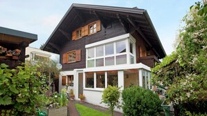 Une maison à deux étages, dotée d’un toit en pente, de grandes fenêtres et d’un petit jardin agrémenté de plantes en pot.